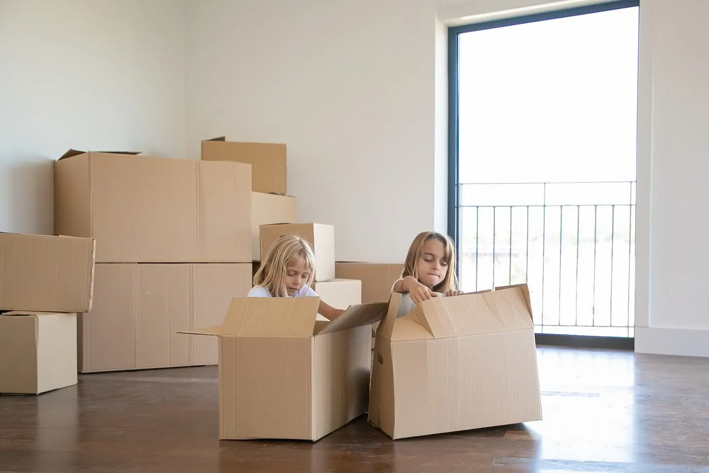 Two cute girls are unpacking belongings in a new apartment, seated on the floor beside open cartoon boxes.