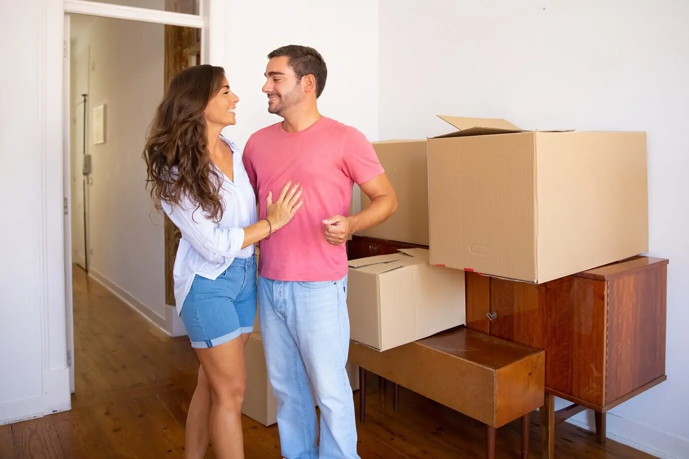 A happy young couple is moving into a new flat, standing beside furniture and carton boxes and discussing unpacking.