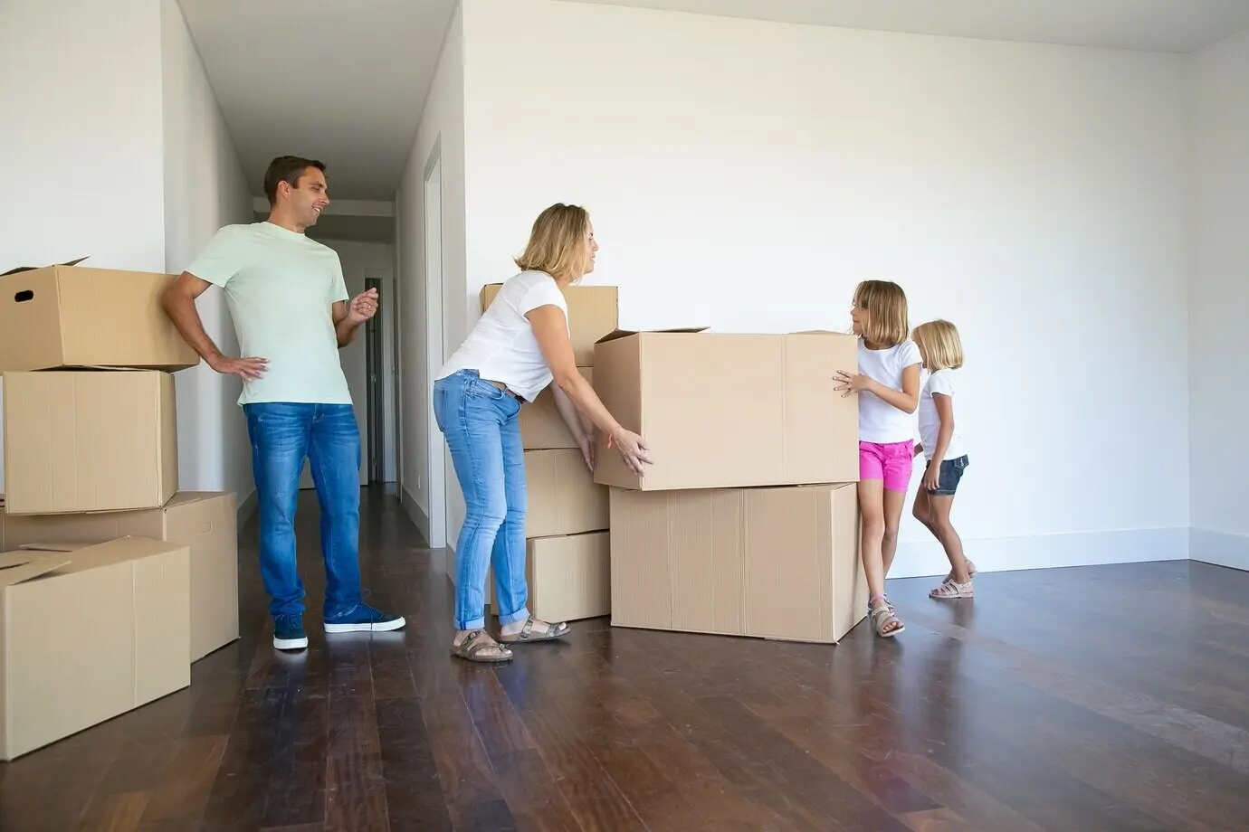 Mom, dad, and two girls carry boxes and stack them in their new empty flat.