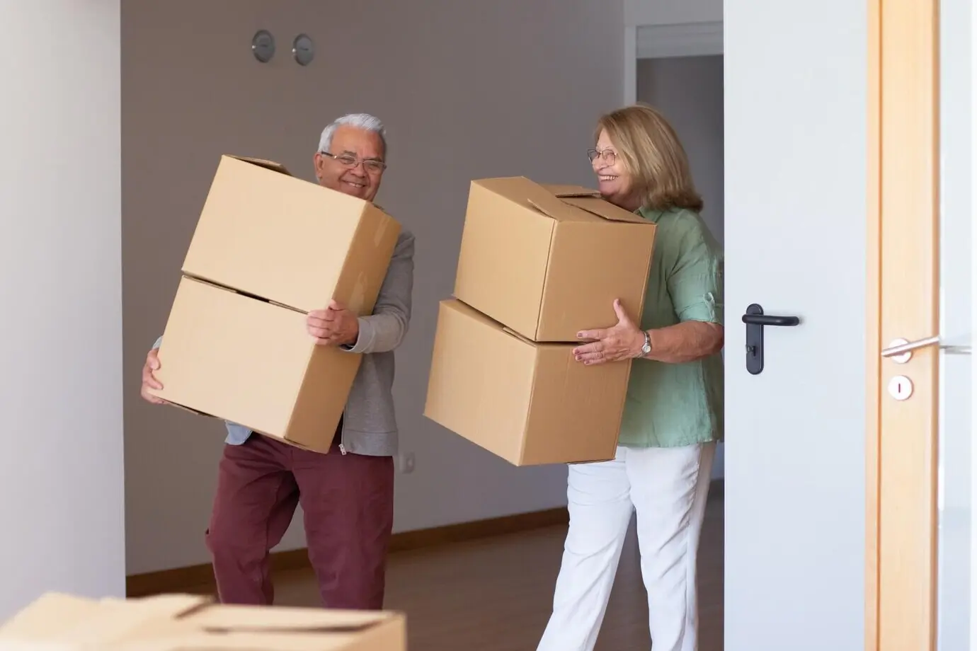 Smiling elderly couple moving into a new house. Retired husband and wife holding cardboard boxes and carrying items. Concept of real estate, purchase, and moving.