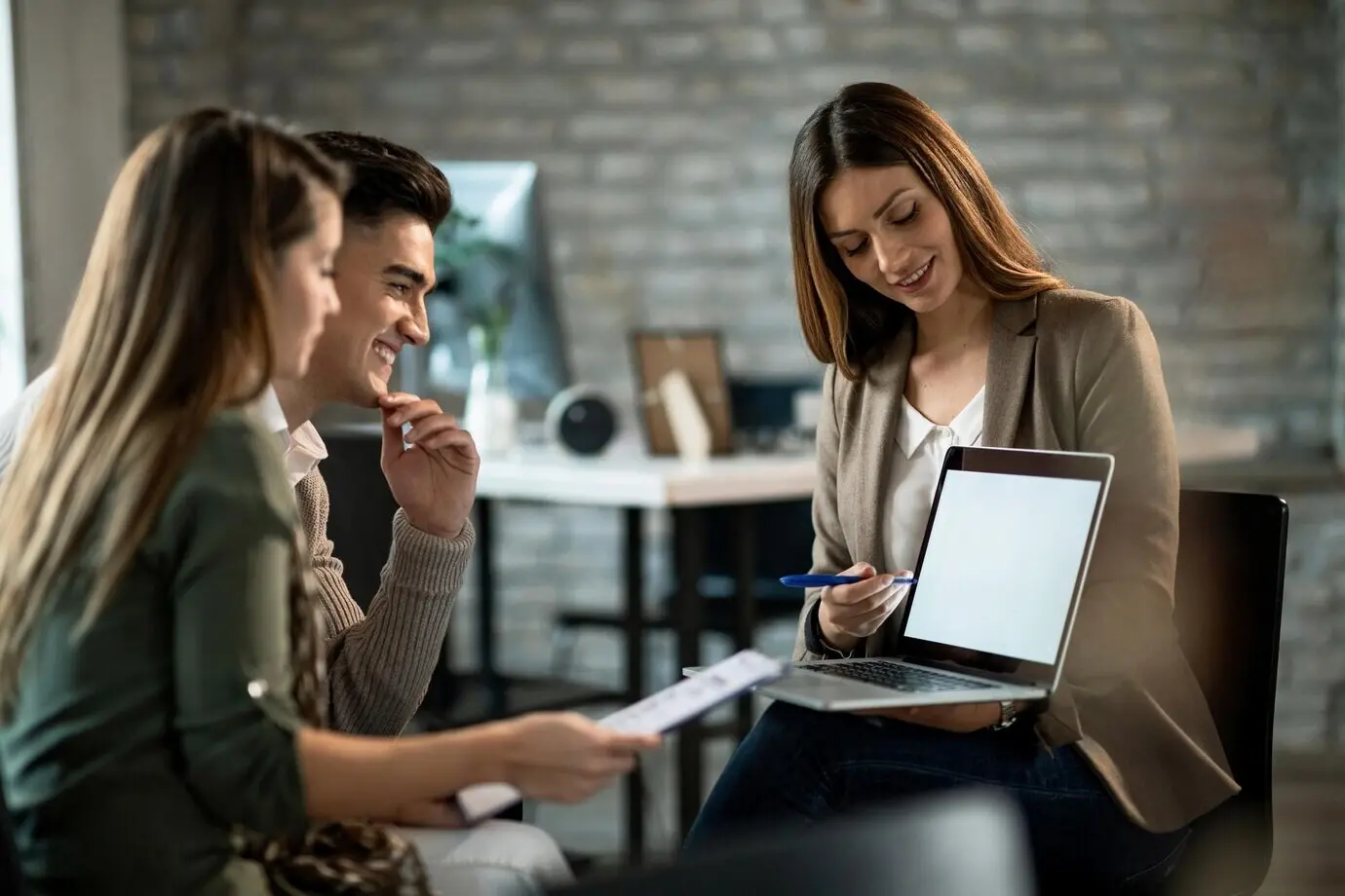 A smiling real estate agent uses a computer during a meeting with a young couple, making investment plans.